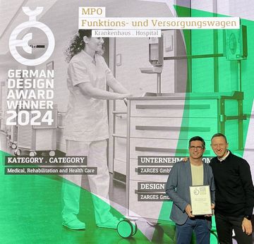 Zwei Mitarbeiter von ZARGES stehen vor einer Leinwand des German Design Award Winner 2024 für MPO Funktions- und Versorgungswagen. Der linke Mitarbeiter hält die Auszeichnung in den Händen.