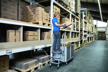 Eine Frau arbeitet im Lager mit Hilfe eines Federbodenwagens, an dem ein Tritt montiert ist.