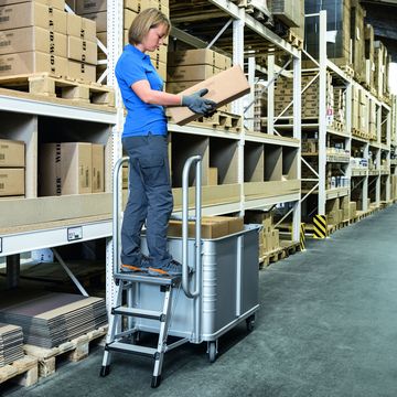 Eine Frau arbeitet im Lager mit Hilfe eines Federbodenwagens, an dem ein Tritt montiert ist.